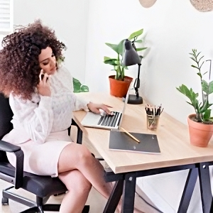 : Spa business coach working in a stylish office, using a laptop and talking on a cell phone. The desk is adorned with a houseplant, flowerpot, and coffee mug.